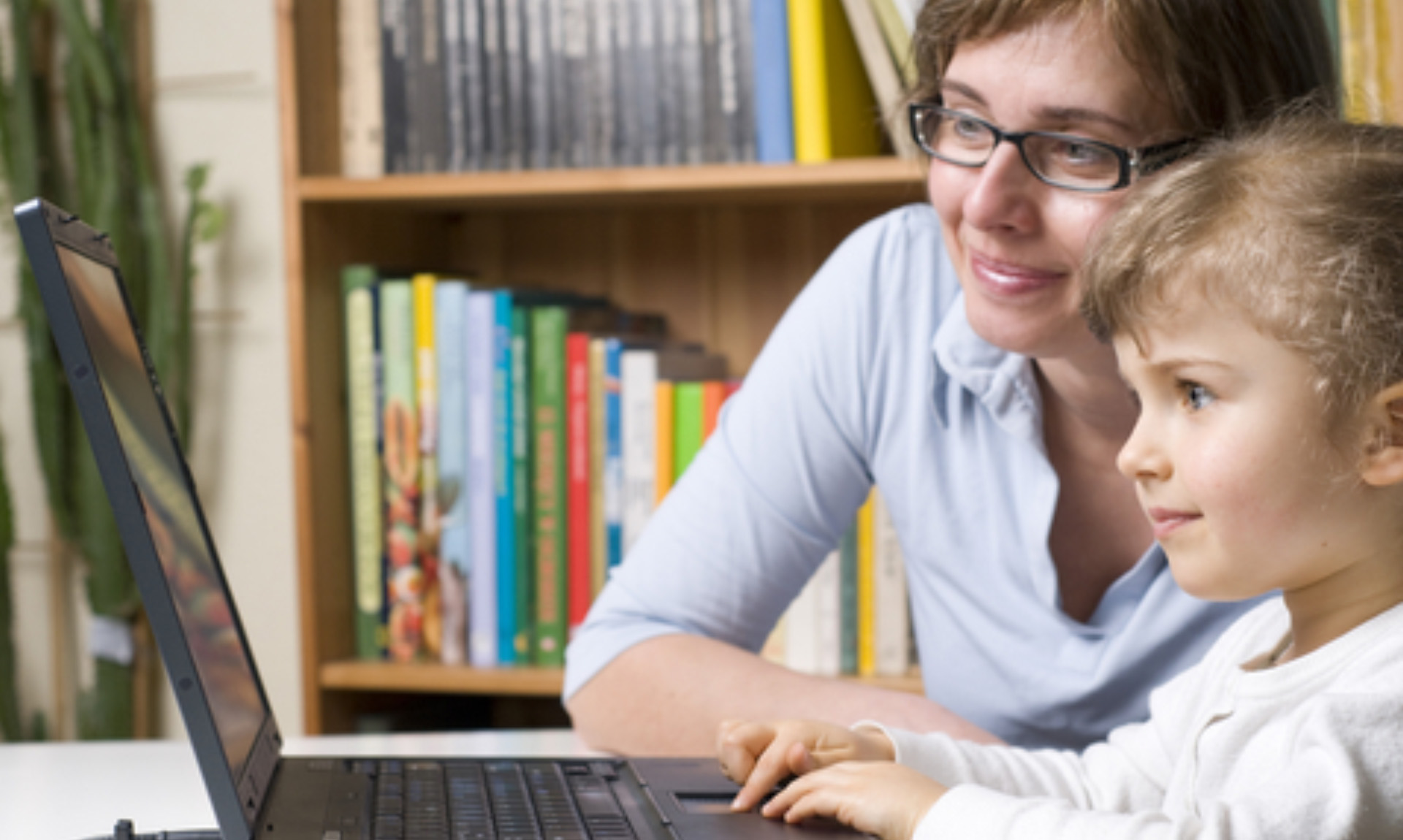 A woman in glasses assists a child with using a laptop.