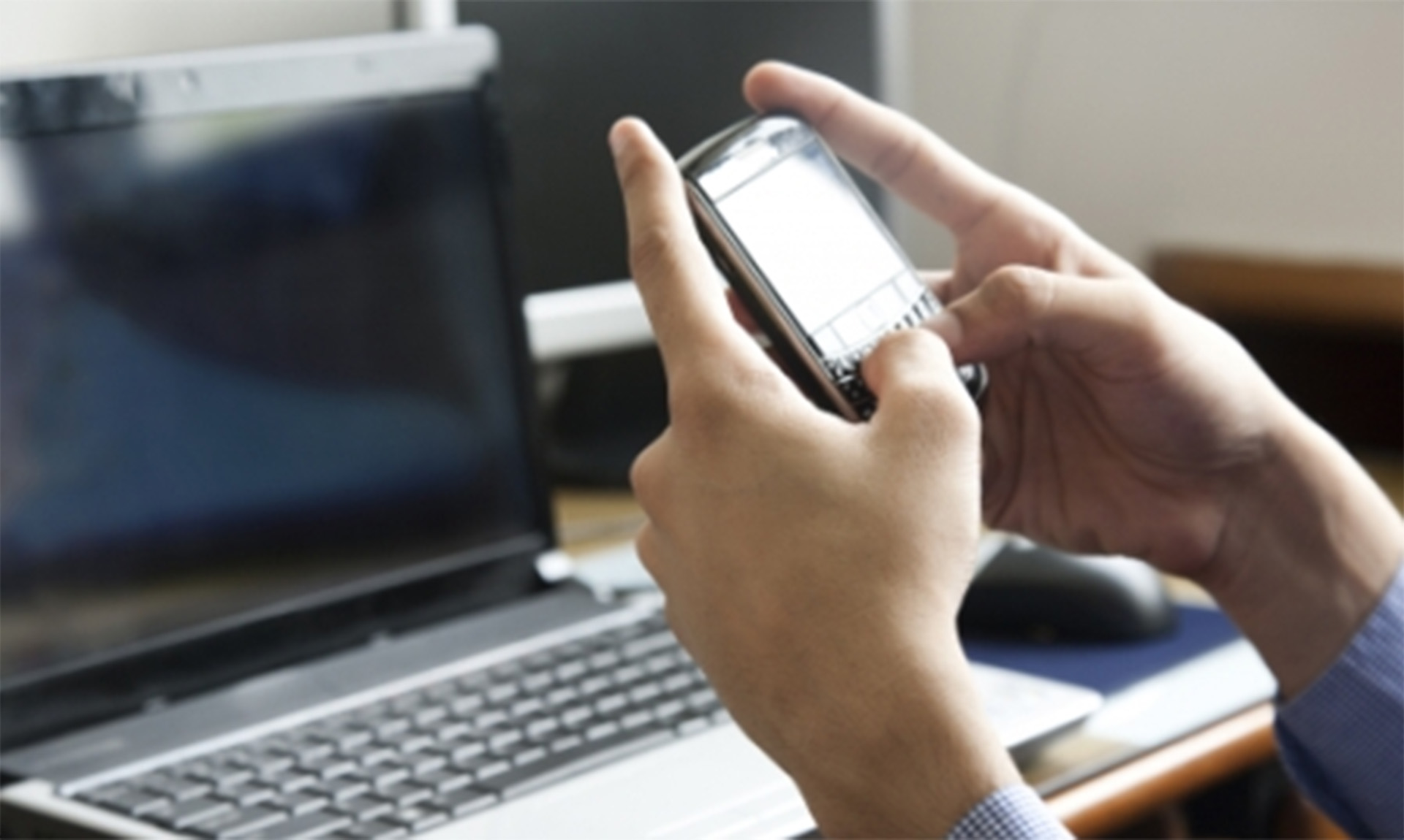 Hands hold a small electronic device with a keyboard. In the background, a desktop computer is visible.