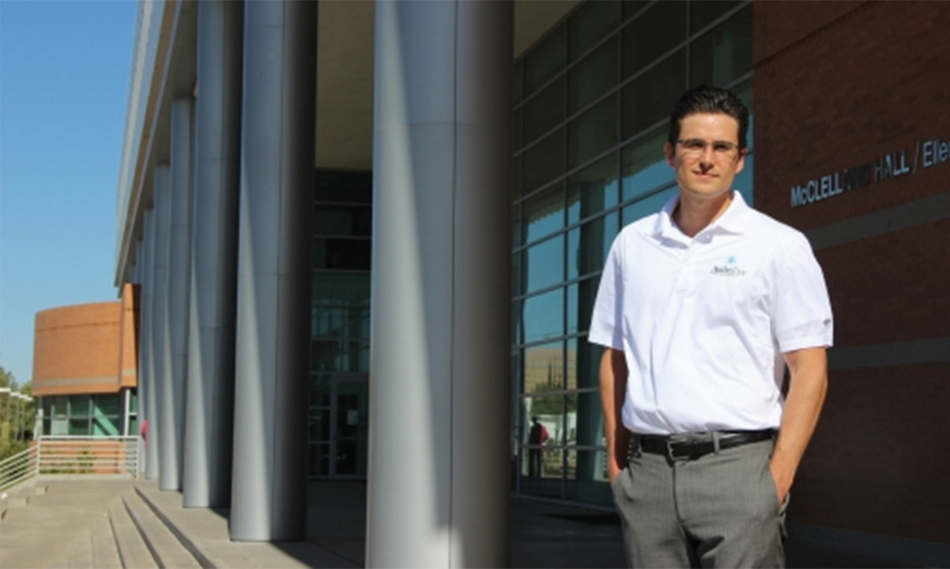Image of a man in a white shirt standing in front of a brick building with silver columns.