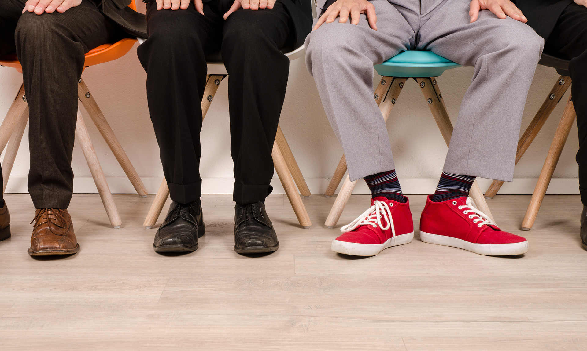 Three people are pictured sitting down. They are visible from the waist down. The person on the right is wearing bright red shoes, in contrast to the business casual attire of the other two people.