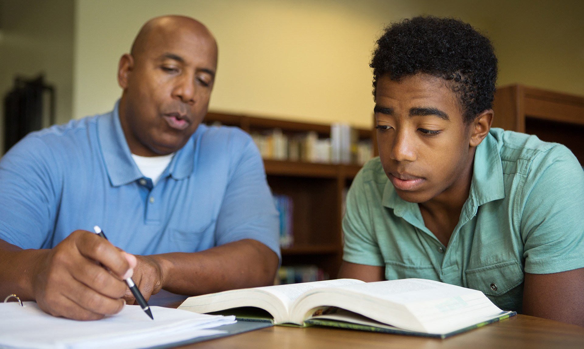 A man and boy sit at a table, looking at an open book and a notebook. The man is pointing to something in the notebook and speaking.