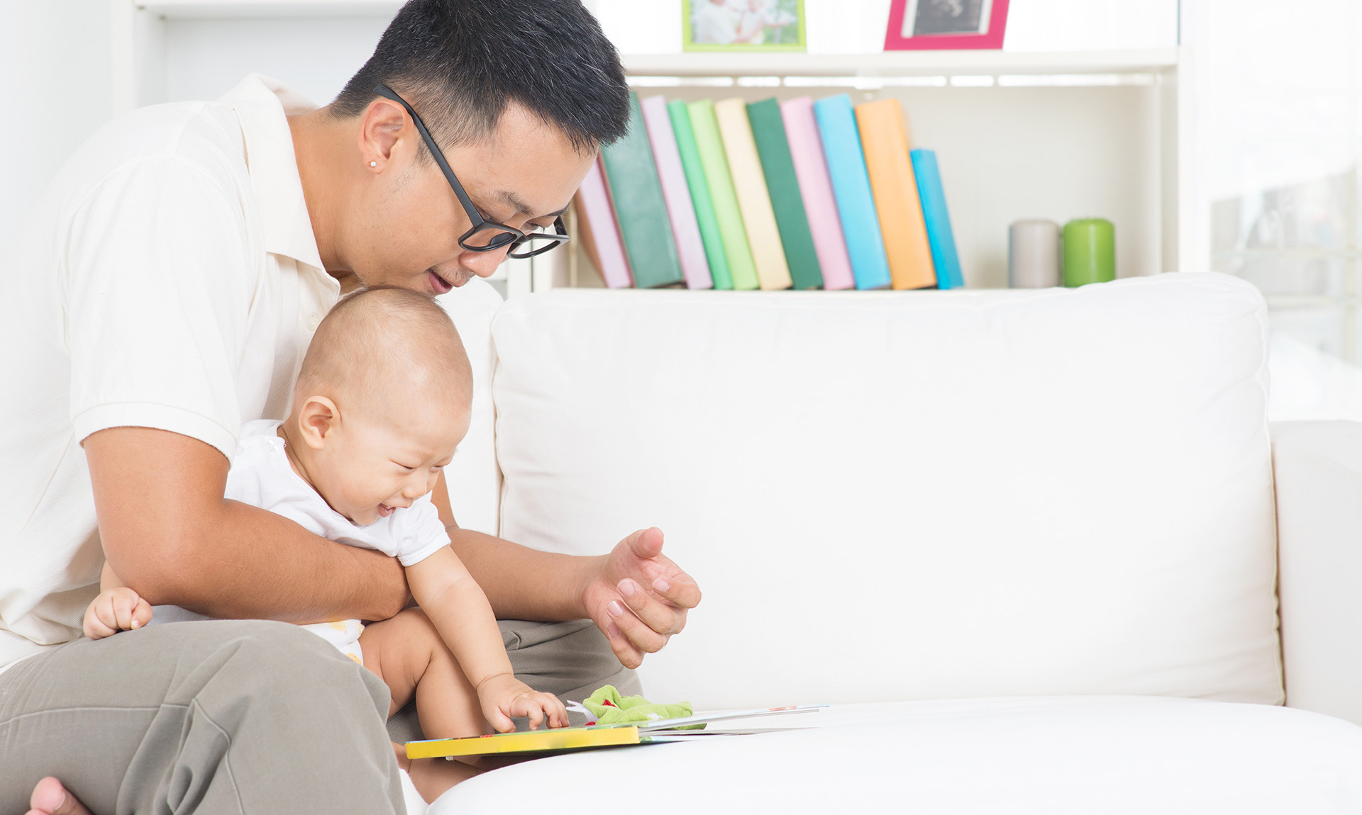 An adult, wearing a white shirt and glasses, holds a small child. Both are engaged with a yellow and green reading toy.