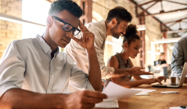 three people working at a wooden table top, closest person is reading a piece of paper in deep contemplation