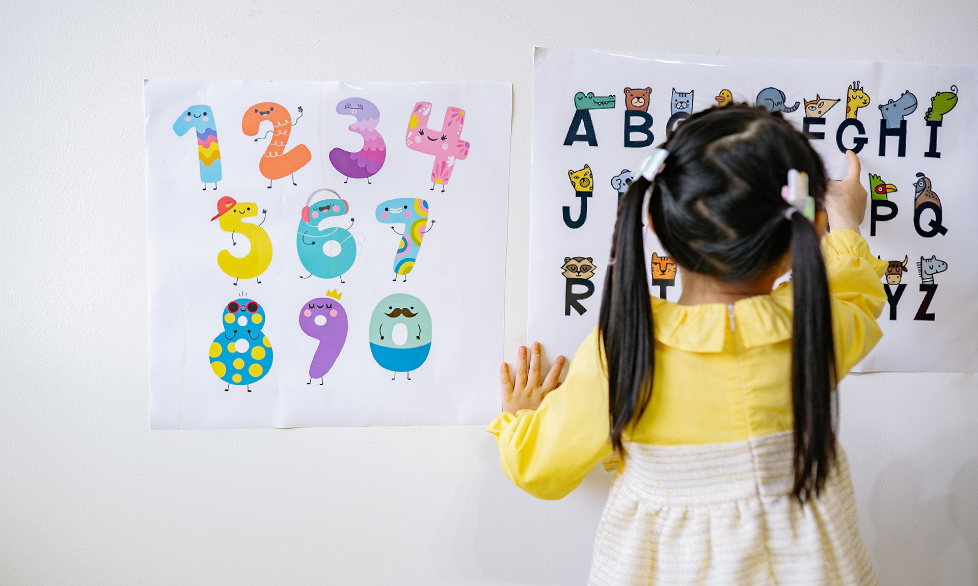 A child with dark hair, pigtails, and a yellow and white dress is visible from the back. She is pointing to a poster on a wall that shows the alphabet illustrated with animals.