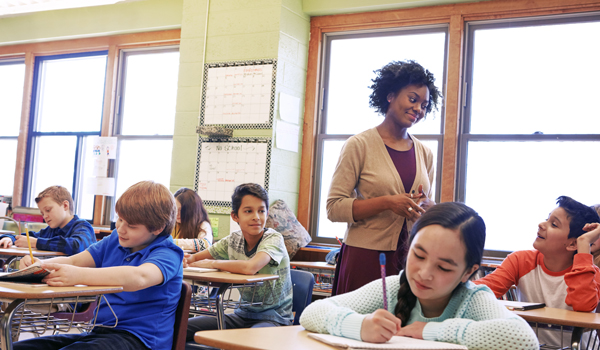 Kids in classroom writing while teacher has a discussion with a student.