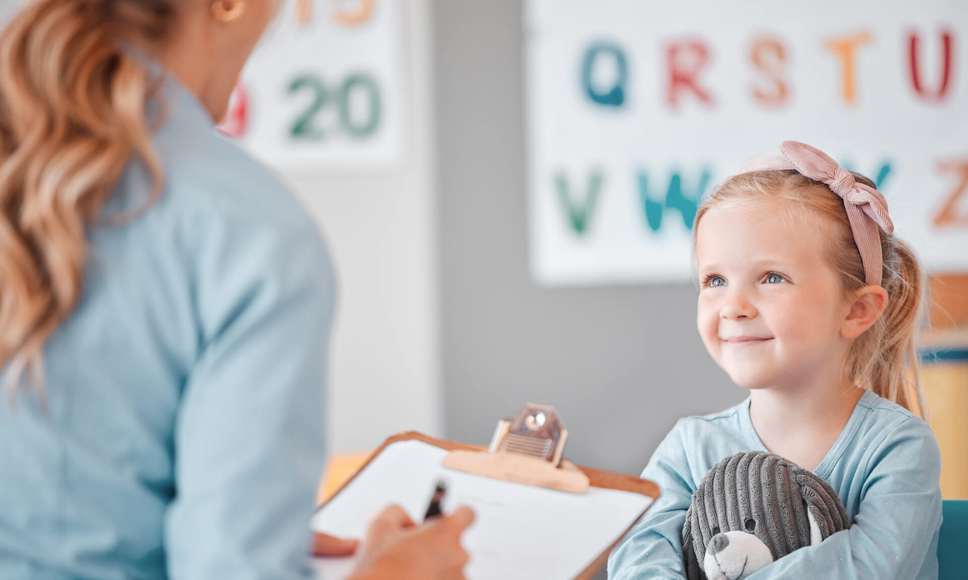 Diagnostic Evaluation Therapist writes notes and smiles, asking a question to a smiling girl hugging a stuffed animal.