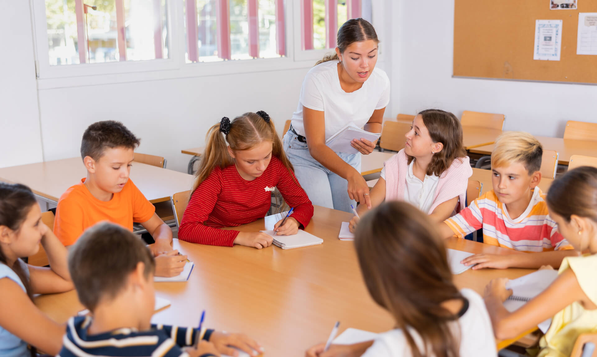 Positive young teacher giving lesson for group of preteen schoolchildren sitting around desk